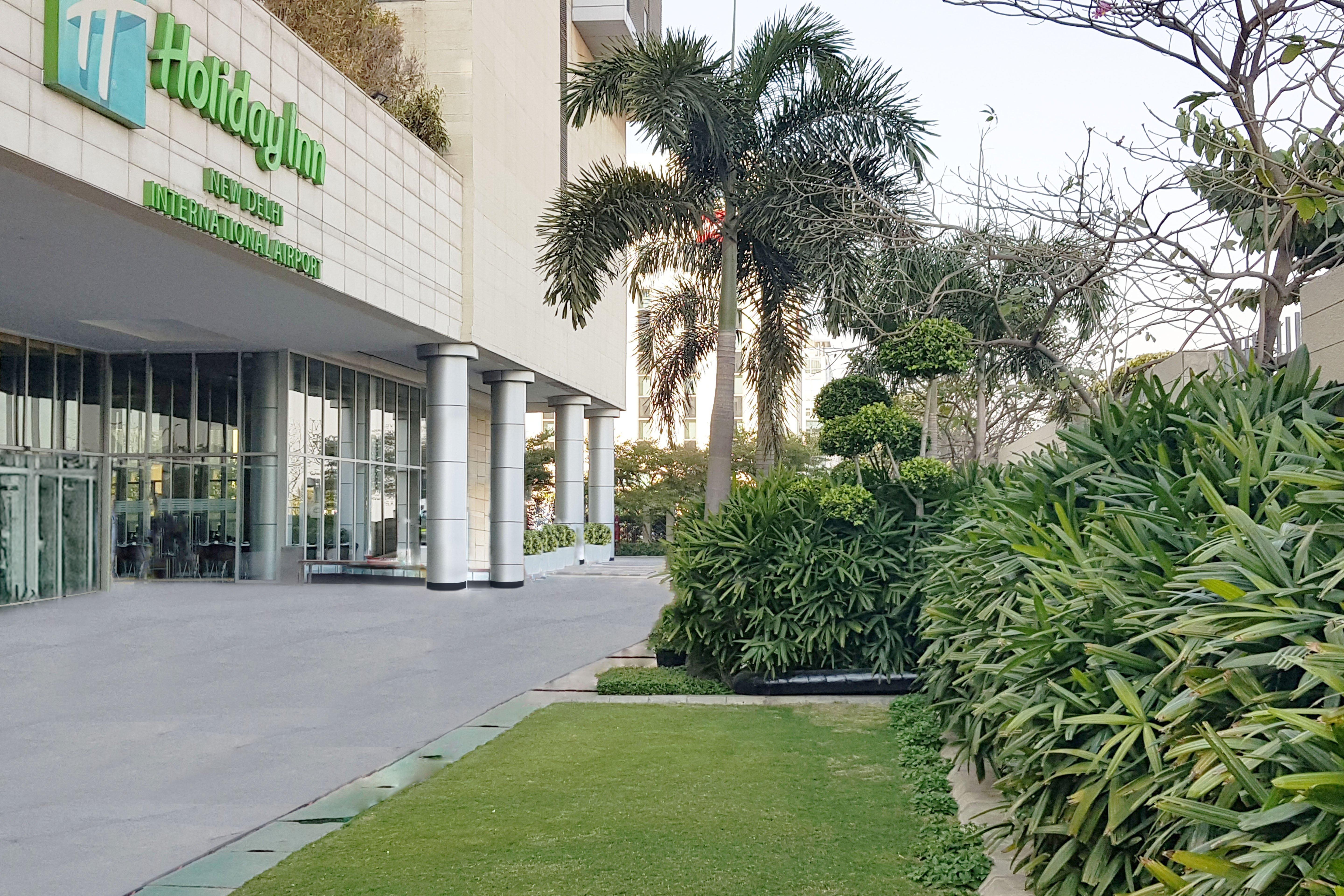 Holiday Inn New Delhi Aerocity By Ihg Экстерьер фото The photo shows the exterior of a Holiday Inn hotel. In the foreground, there is a well-maintained green lawn and various ornamental plants, creating a pleasant garden area. The building features large glass doors and columns, and the hotel's name an